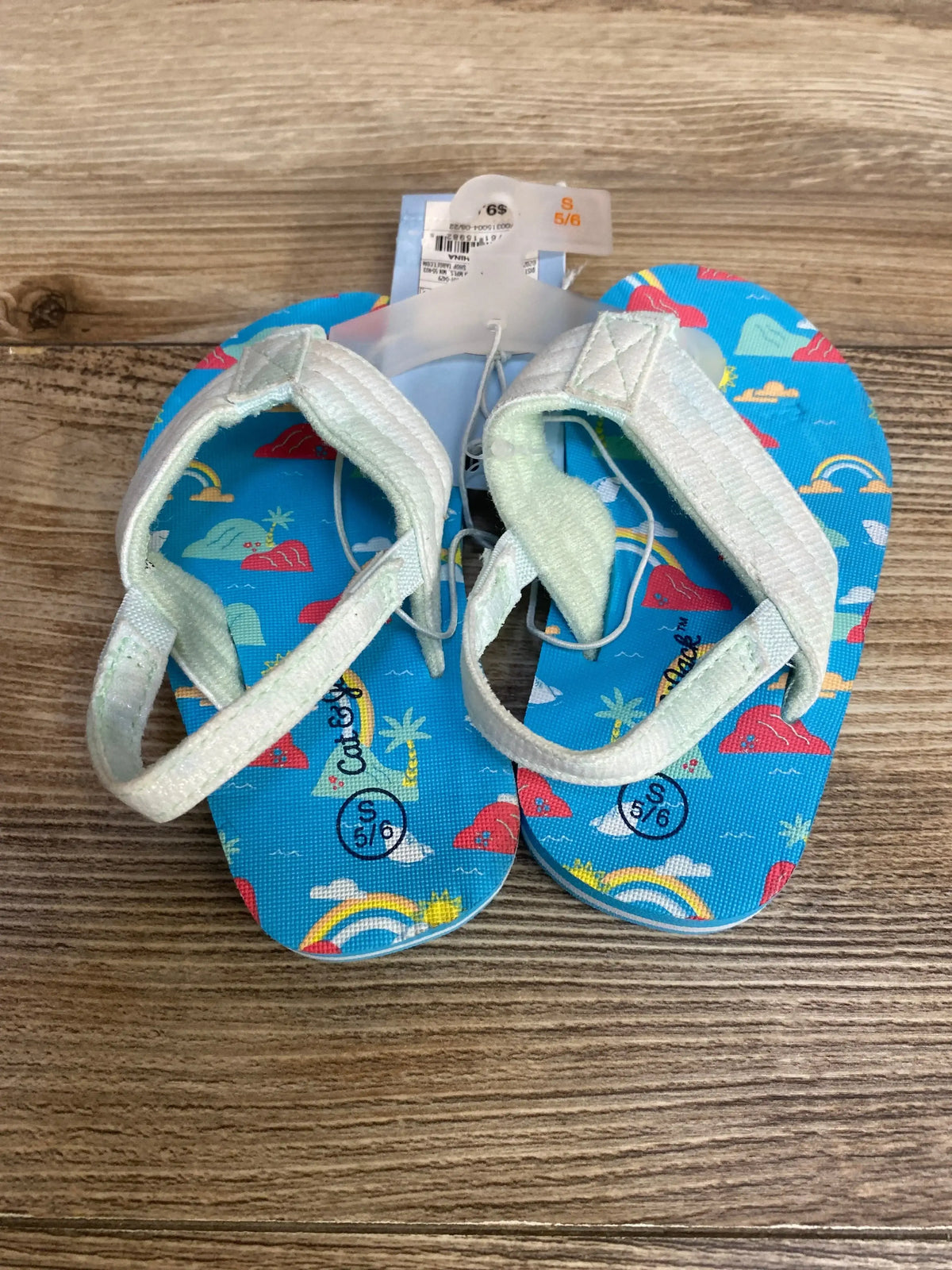 Top view of Cat & Jack toddler sandals with blue rainbow print footbeds, white straps, size 5/6 label on wood floor