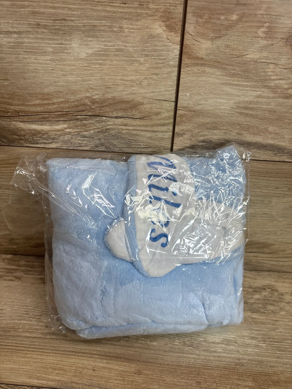 Folded light blue baby hooded towel with bunny ears and white trim, plush fabric, wrapped in clear plastic on wood