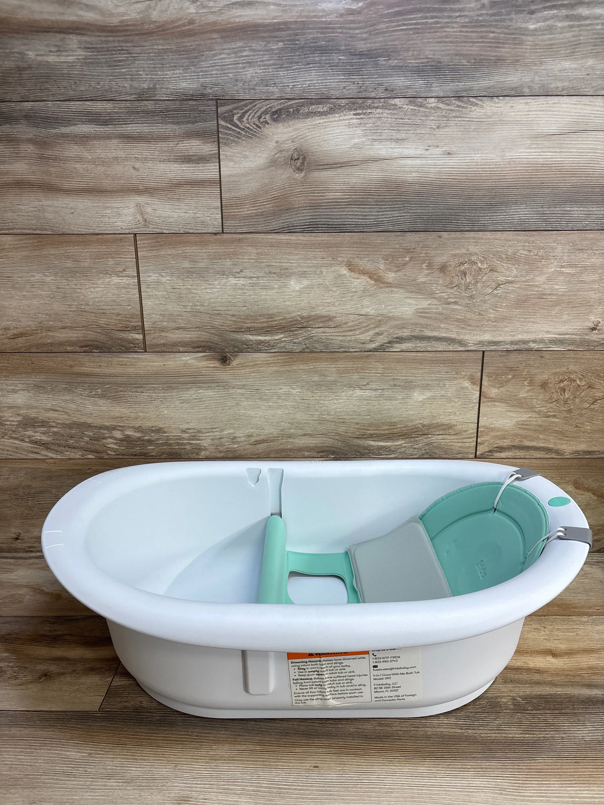 Side view of Frida Baby 4-in-1 Grow-With-Me Bath Tub showing white tub with mint-green sling and infant support on wood floor