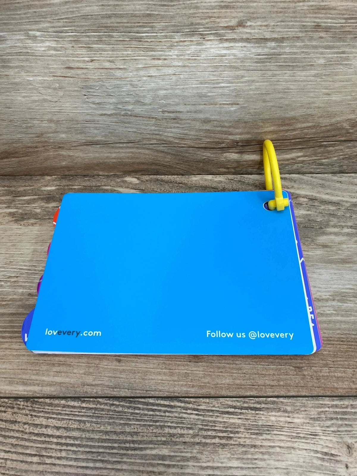 Top-down view of Lovevery The Babbler play guide (orange, 13-15 months) showing blue back cover and yellow plastic ring on wooden surface