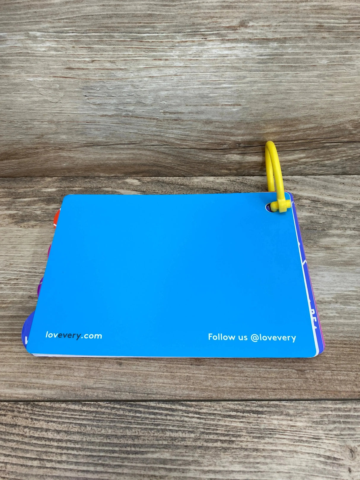 Top-down view of Lovevery The Babbler play guide (orange, 13-15 months) showing blue back cover and yellow plastic ring on wooden surface
