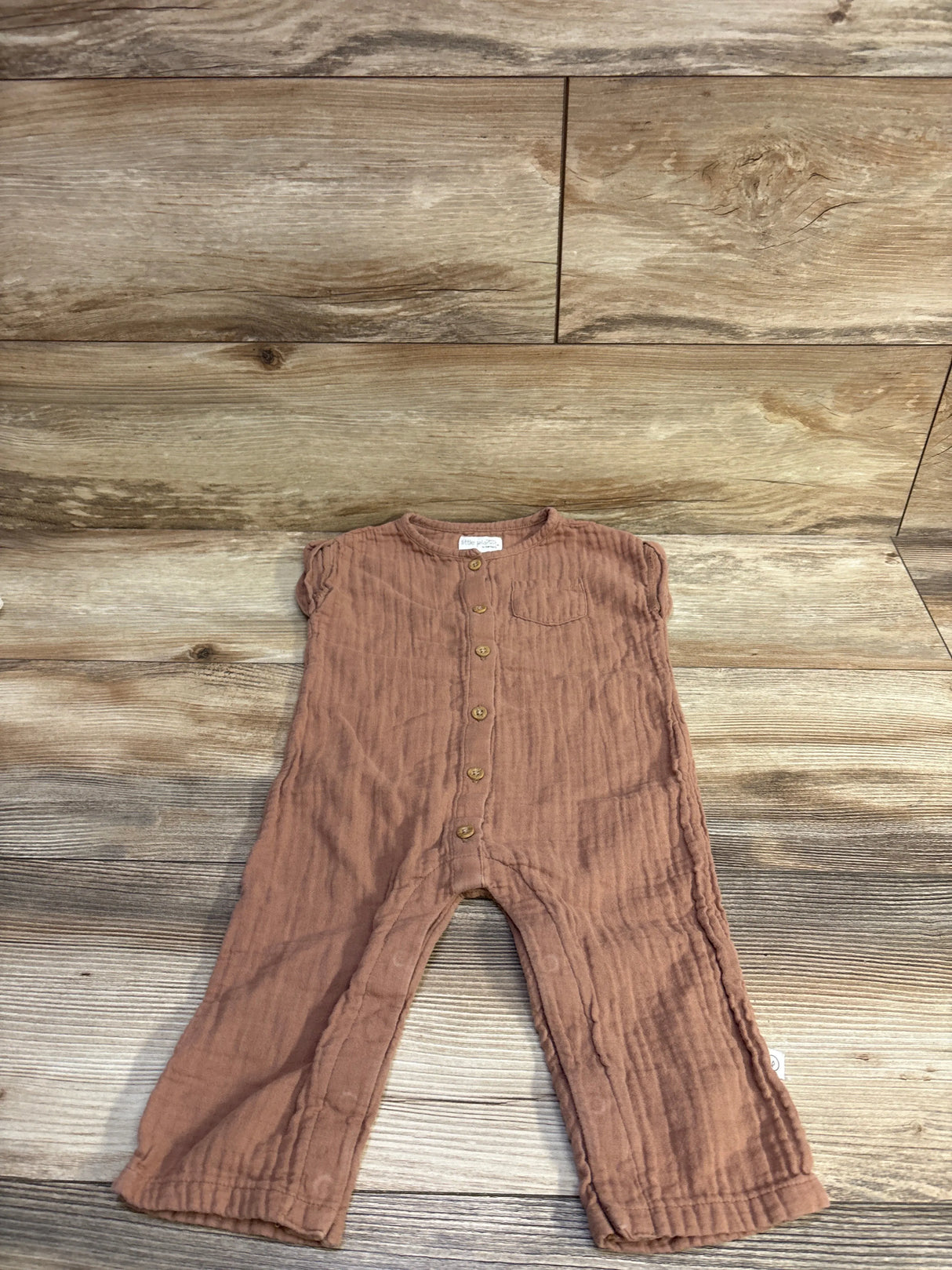 Flat lay of Little Planet brown gauze button-front jumpsuit, size 12 months, showing wooden buttons and cap sleeves on wood background