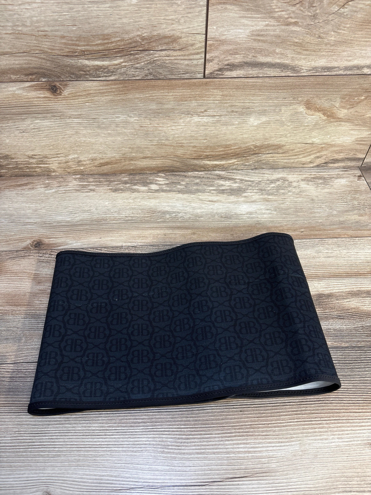 Folded flat lay of Belly Bandit postpartum belly wrap showing black fabric with subtle repeating BB monogram on wood background
