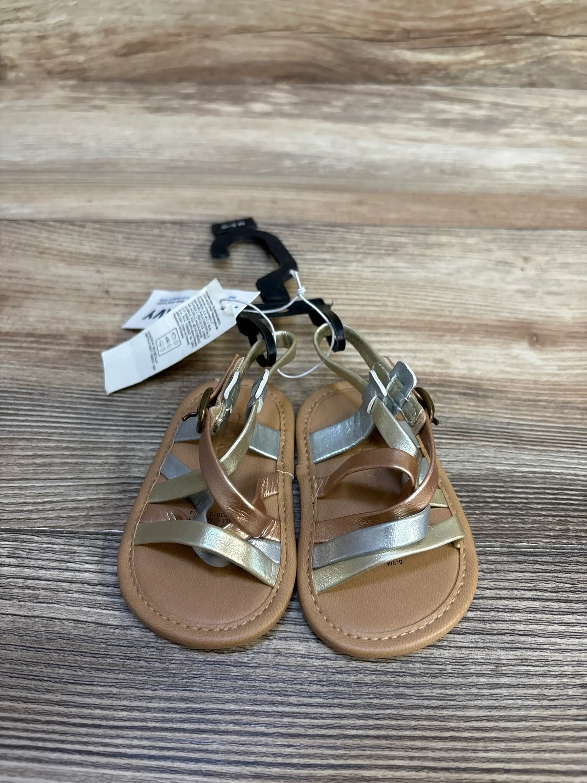 Top view of Old Navy metallic baby sandals size 1/2C (0-3M) with silver and rose-gold crisscross straps and ankle buckles on a wood surface