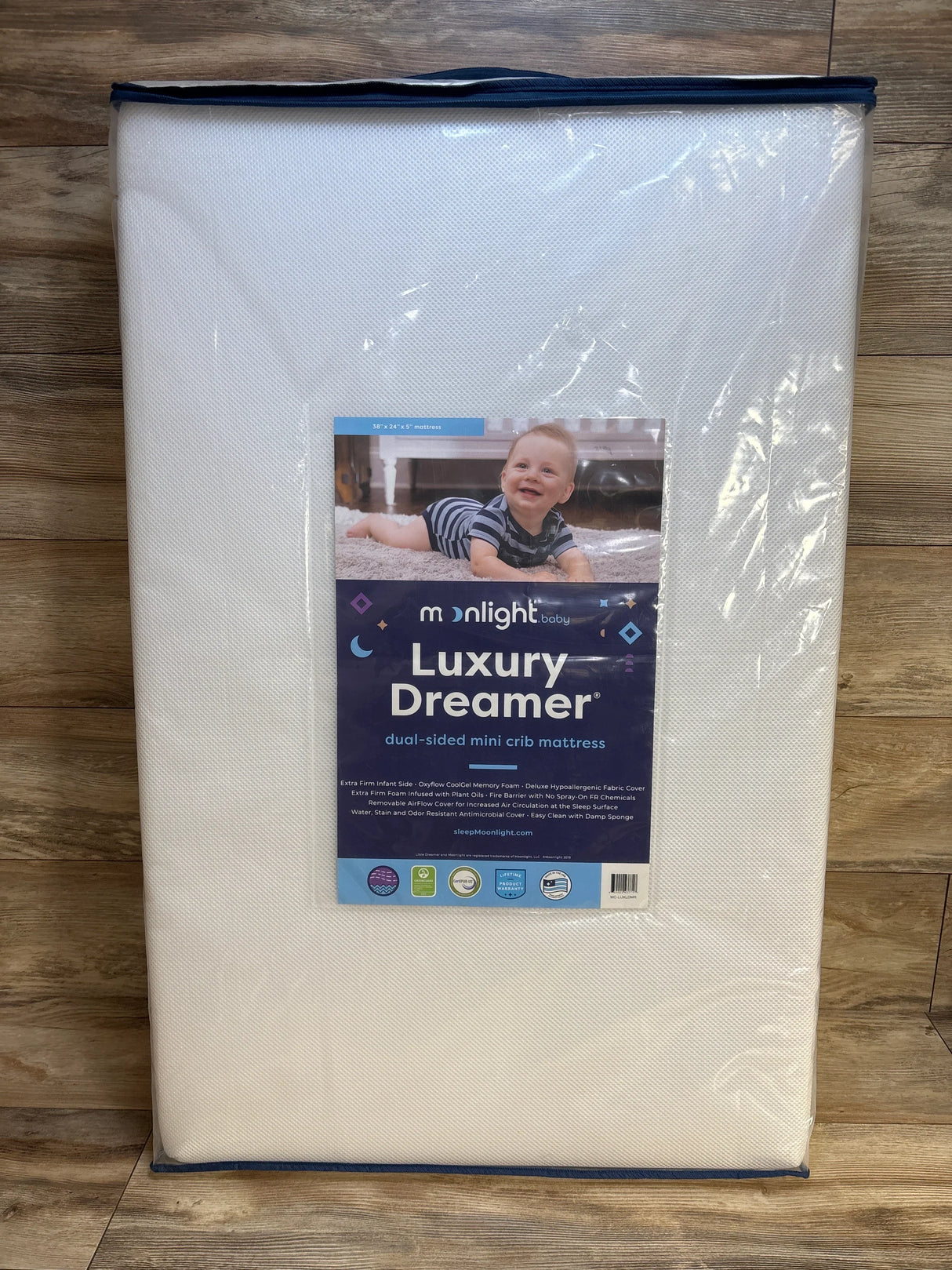 Front view of Moonlight Slumber Luxury Dreamer mini crib mattress showing dual-sided design and product label with dimensions 38 x 24 x 5 in on wood floor