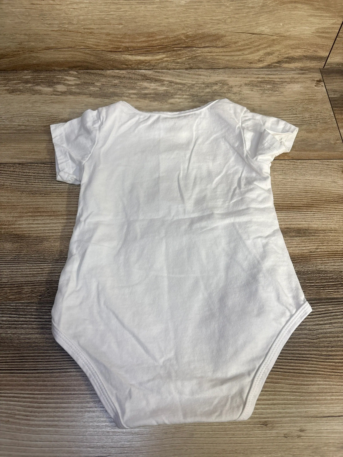 Front view of white 1st birthday bodysuit in size 12 months showcasing short sleeves and rounded neckline on a woodgrain background