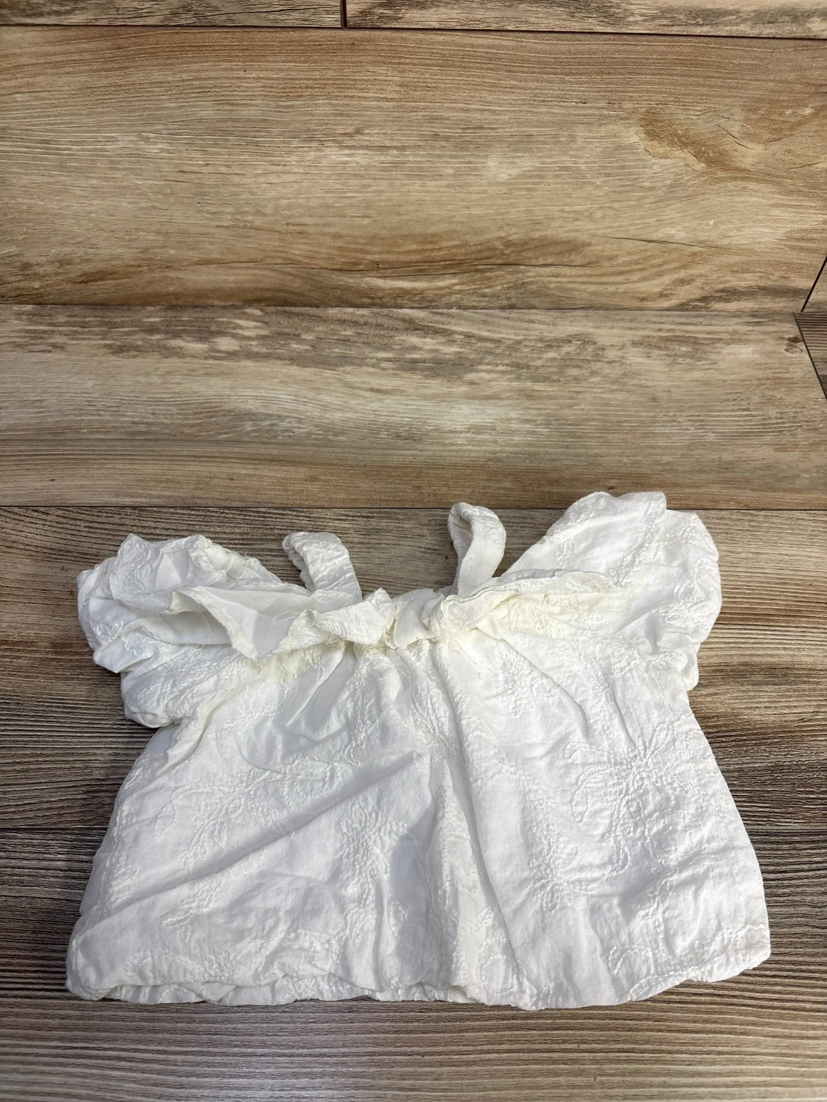 Flat lay of white baby cold-shoulder top showcasing floral embroidery and ruffled sleeves on wood background