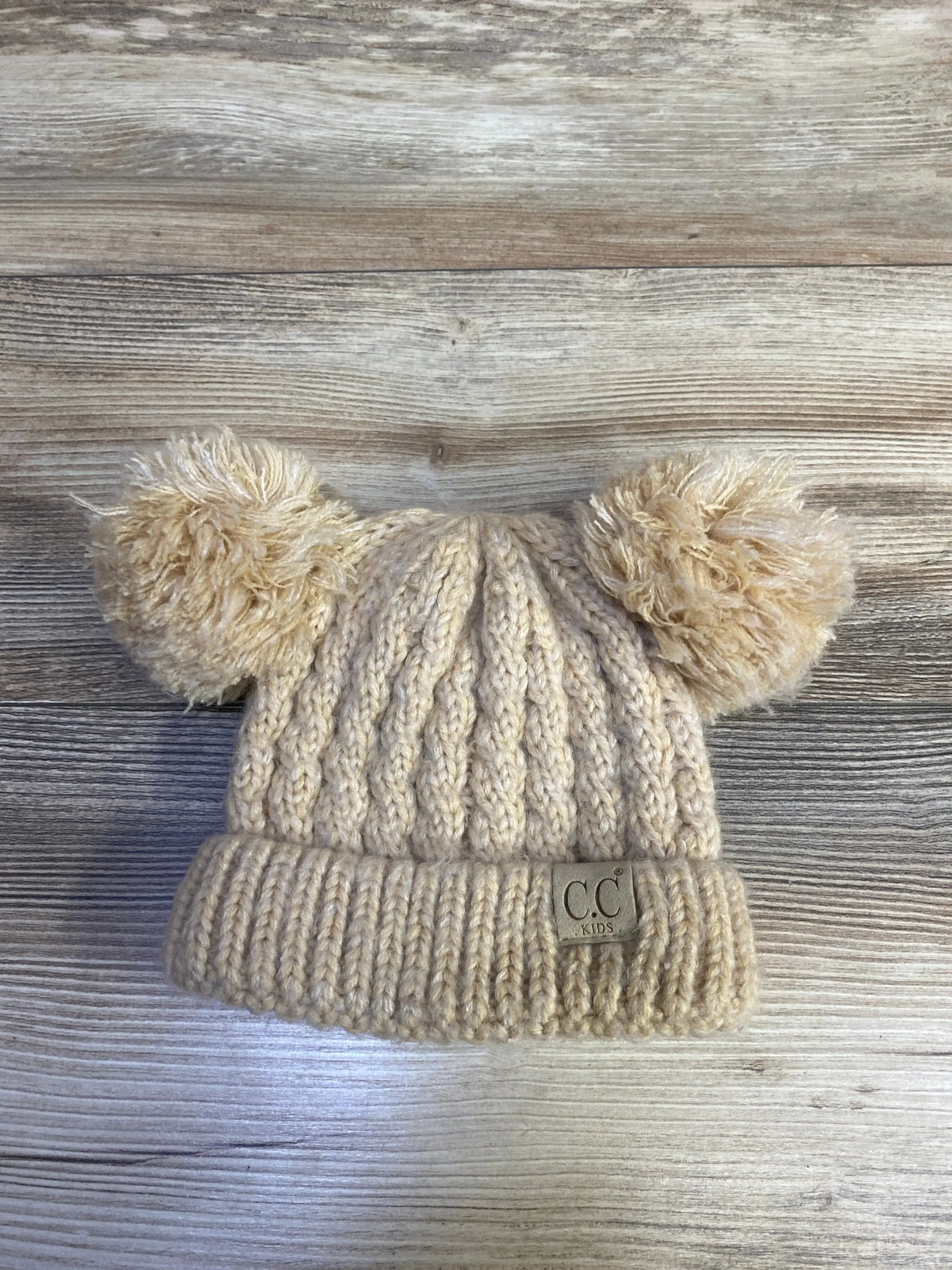 Front view of C.C Kids beige chunky knit pom-pom beanie showcasing two pom-poms and ribbed texture on wood grain