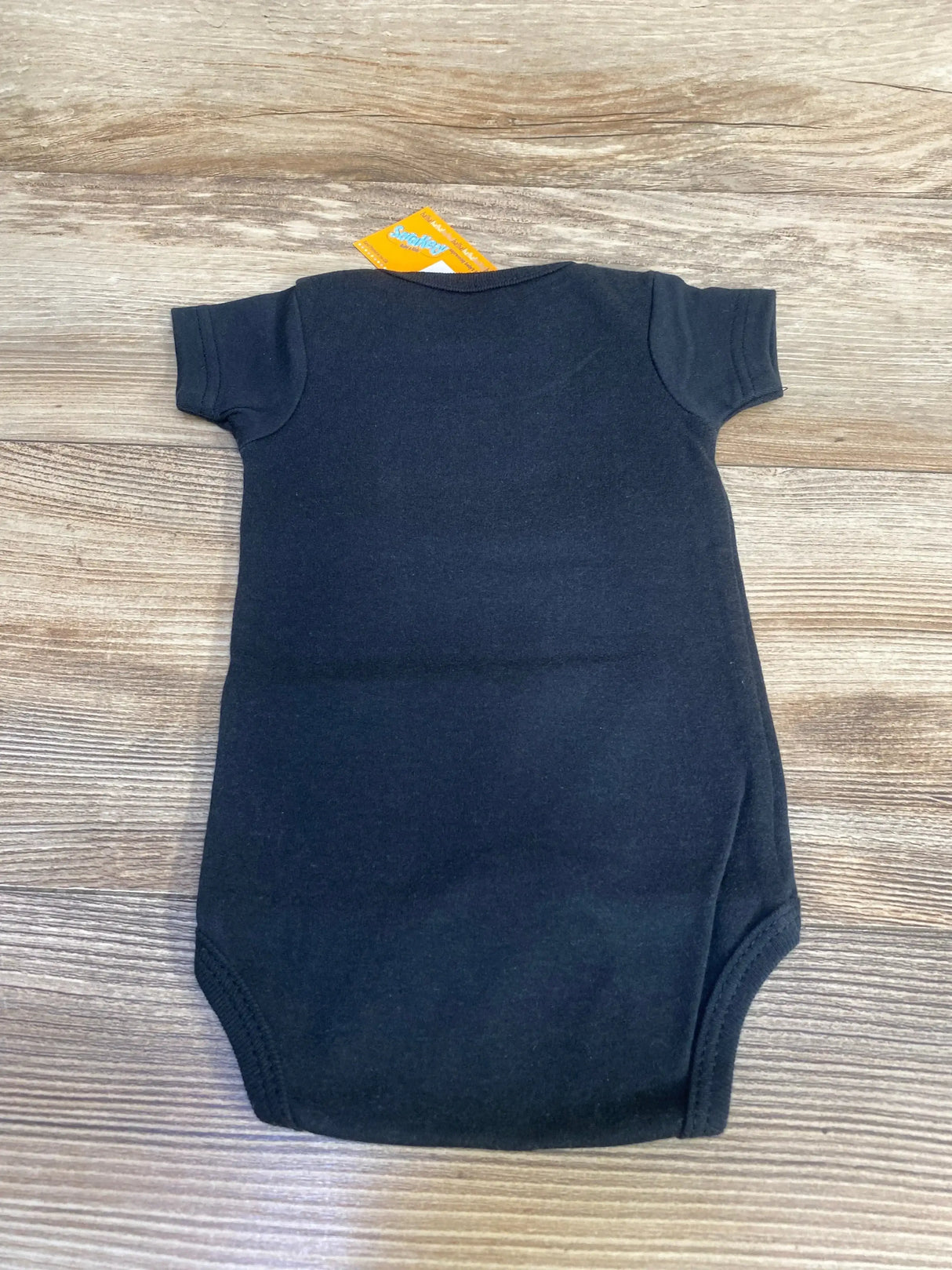 Back view of black baby bodysuit size 0-6 months showcasing short sleeves and envelope neckline with tag, laid flat on wood background