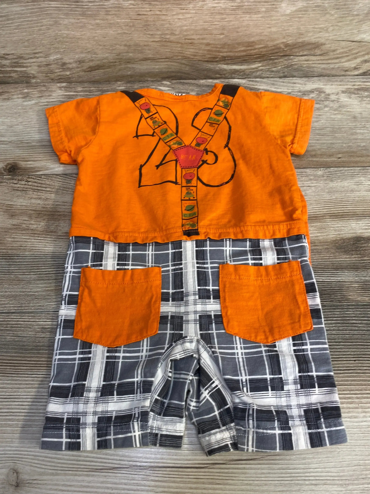 Front flat lay of orange baby romper with printed suspenders and gray plaid shorts with orange pockets on wood background