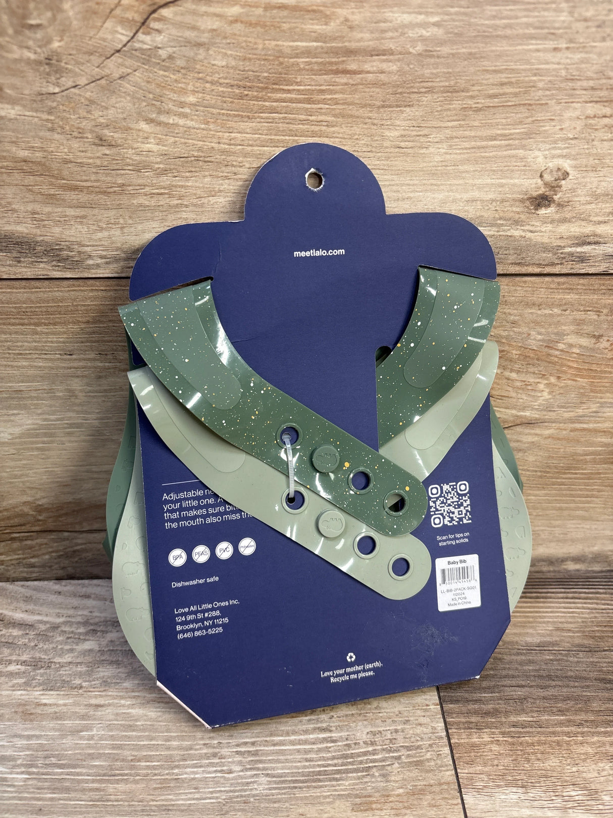 Front view of two Lalo silicone baby bibs in sage with speckled finish and adjustable snap closures on navy backing against wood background