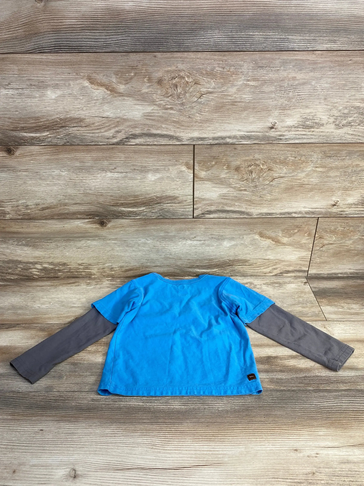 Back view of TEA robot layered tee in blue showcasing gray long sleeves and brand tag on wood plank floor