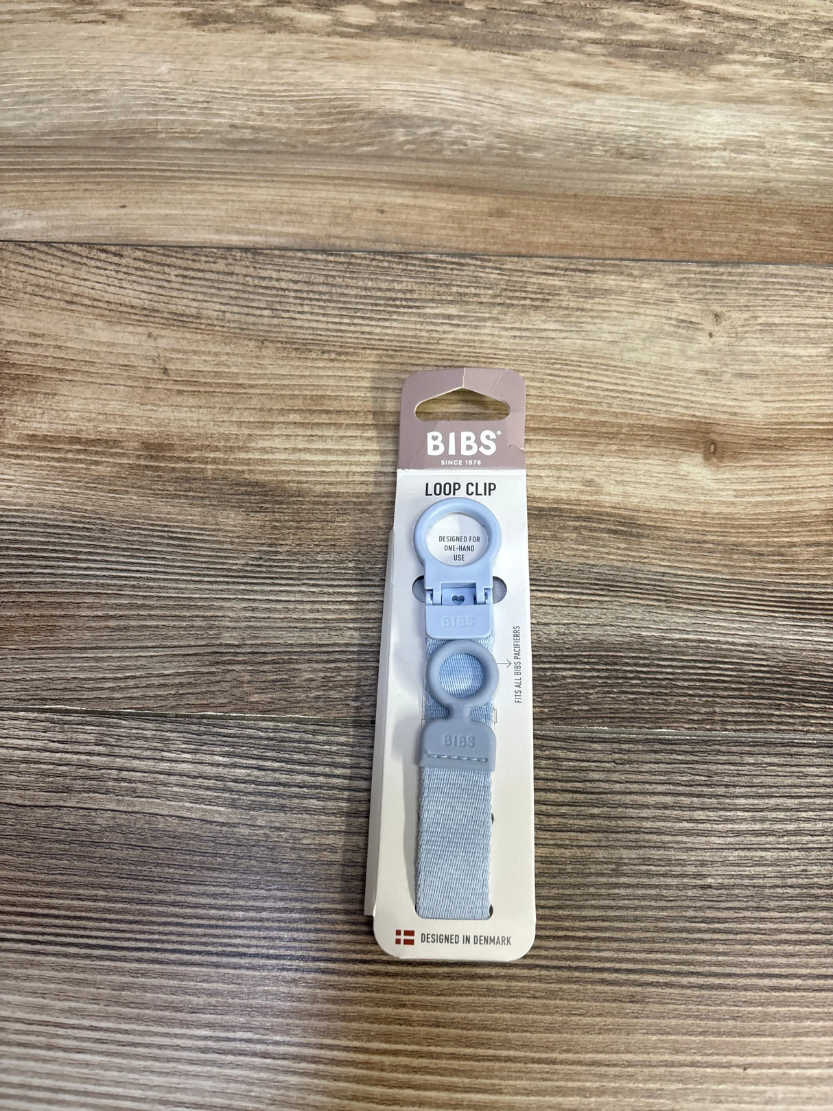 Top-down view of BIBS Loop Clip pacifier holder in light blue on card packaging showing clip and strap on wood background