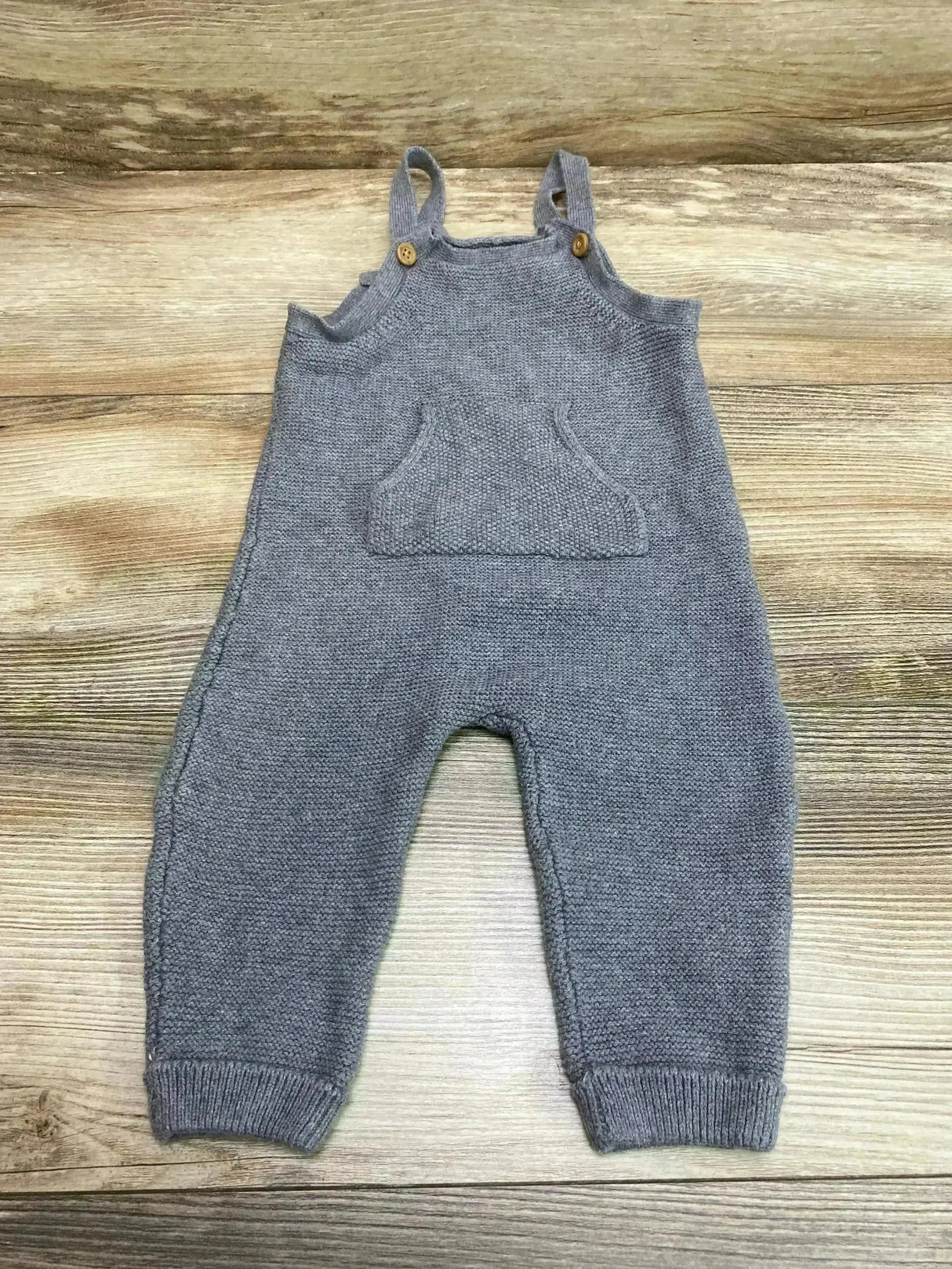 Flat lay of grey knit baby overalls showcasing front kangaroo pocket, button shoulder straps, and ribbed ankle cuffs with wood plank background