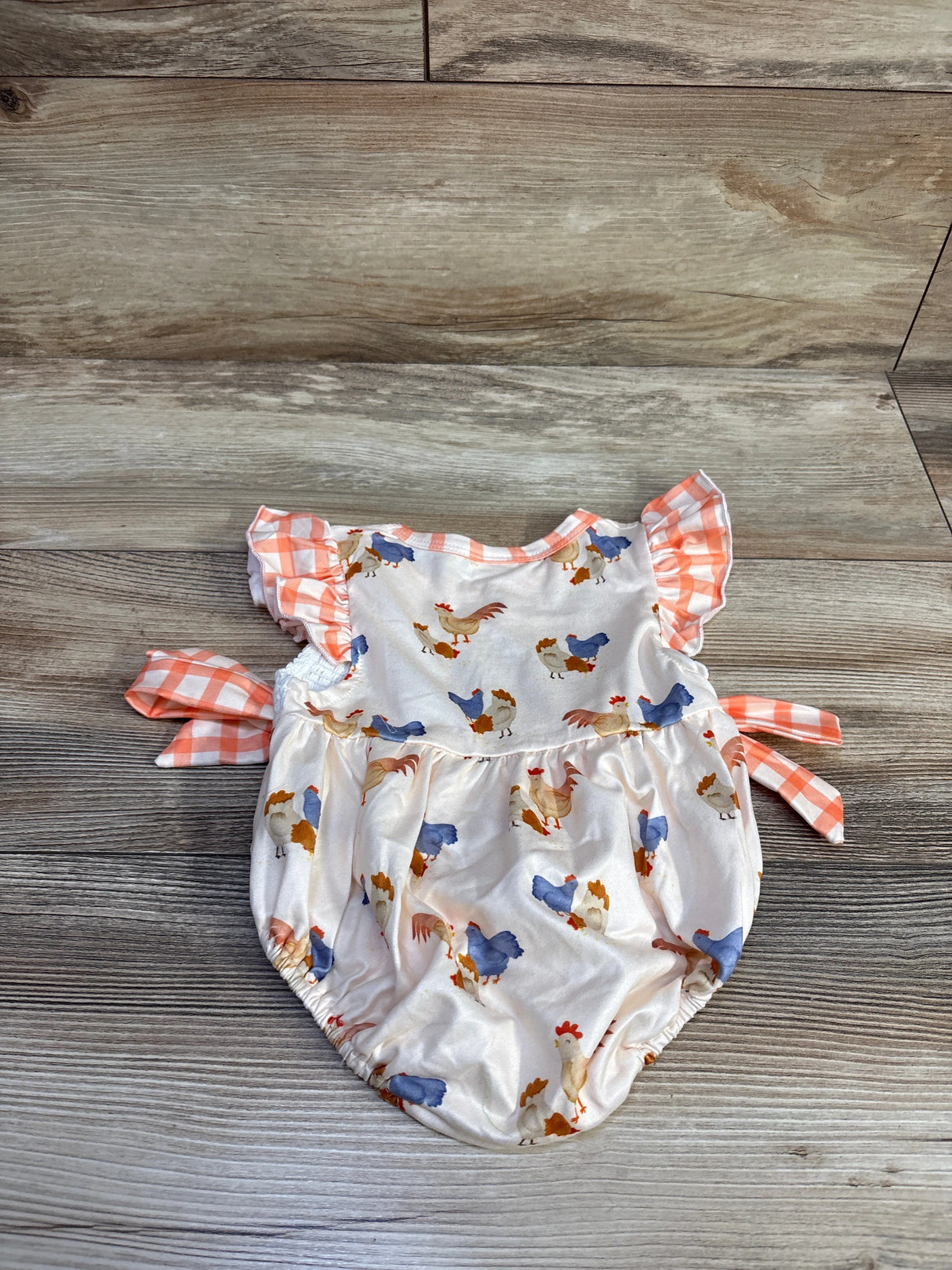Flat lay of ivory baby bubble romper with smocked yoke, rooster print and orange gingham ruffle sleeves on wood background