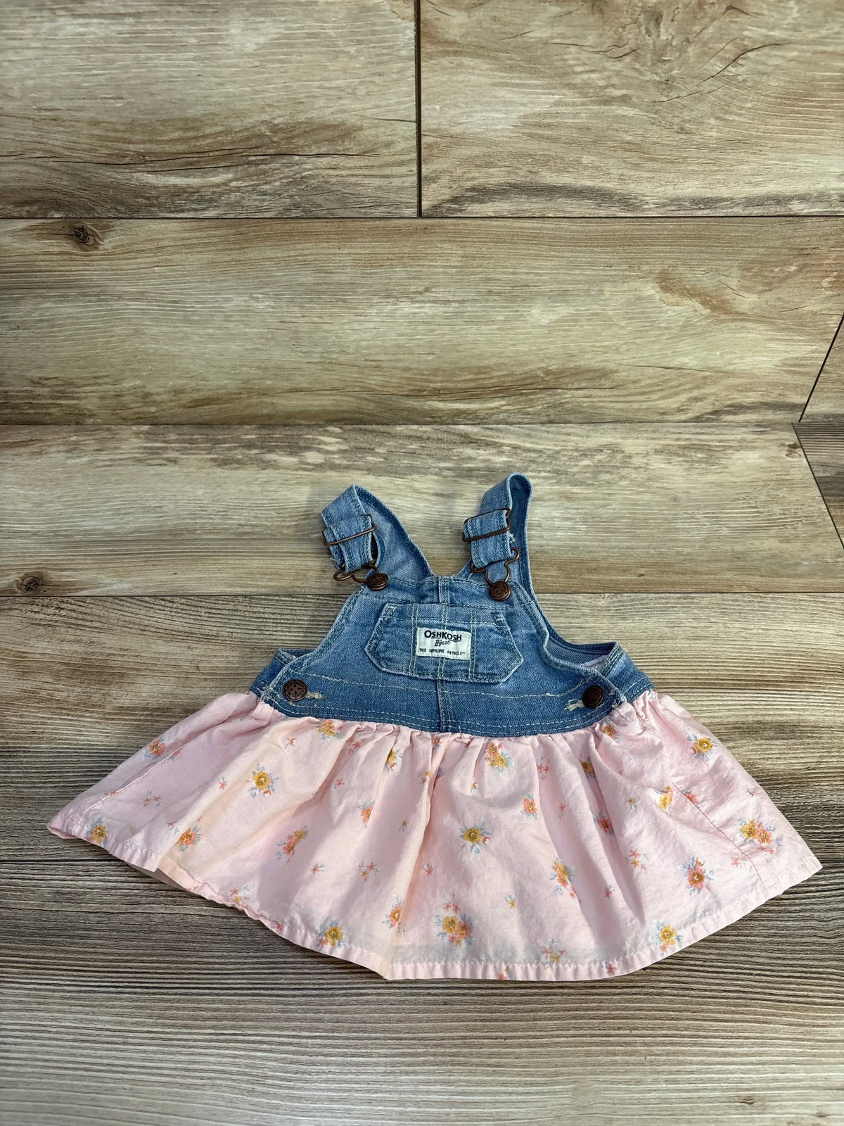 Flat lay of Oshkosh denim skirtall showcasing pink floral skirt, adjustable bib straps and front pocket on wood plank background