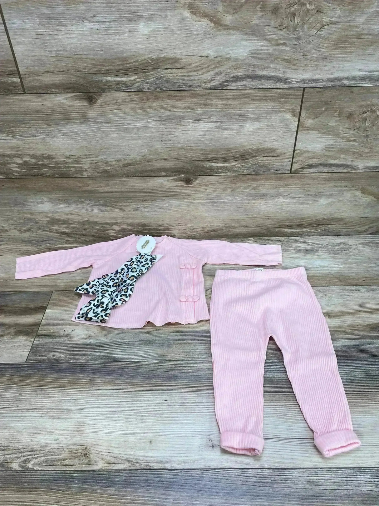 Flat lay of Mud Pie 3-piece ribbed pink set: bow-accent top, matching pants, and leopard-print scarf on wood floor