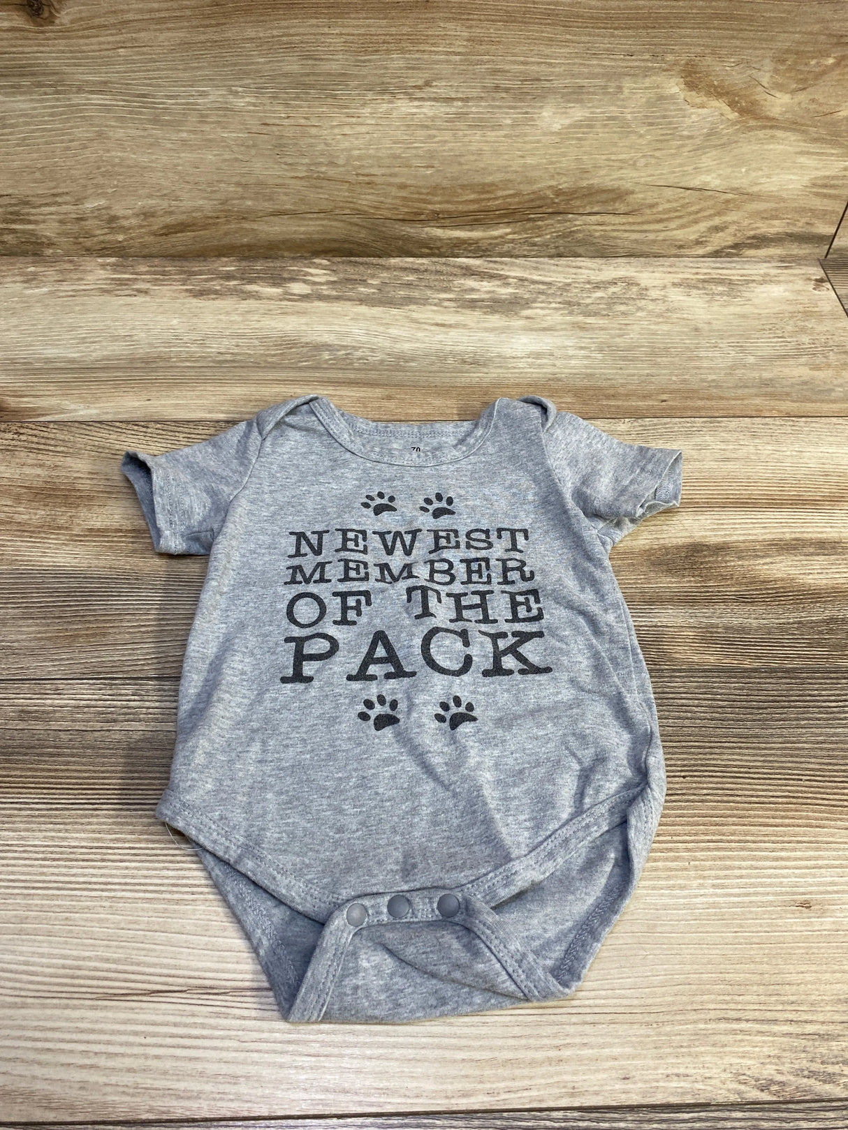 Top-down view of grey baby bodysuit (3-6 months) showcasing ‘NEWEST MEMBER OF THE PACK’ printed text and paw print graphics on wood background