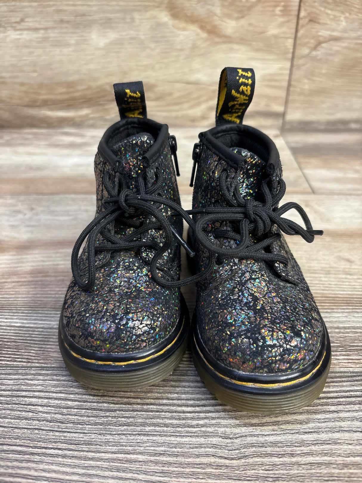 Front view of Dr. Martens 1460 Crinkle Metallic boots showing black glittered upper, yellow welt stitch, clear sole on wood