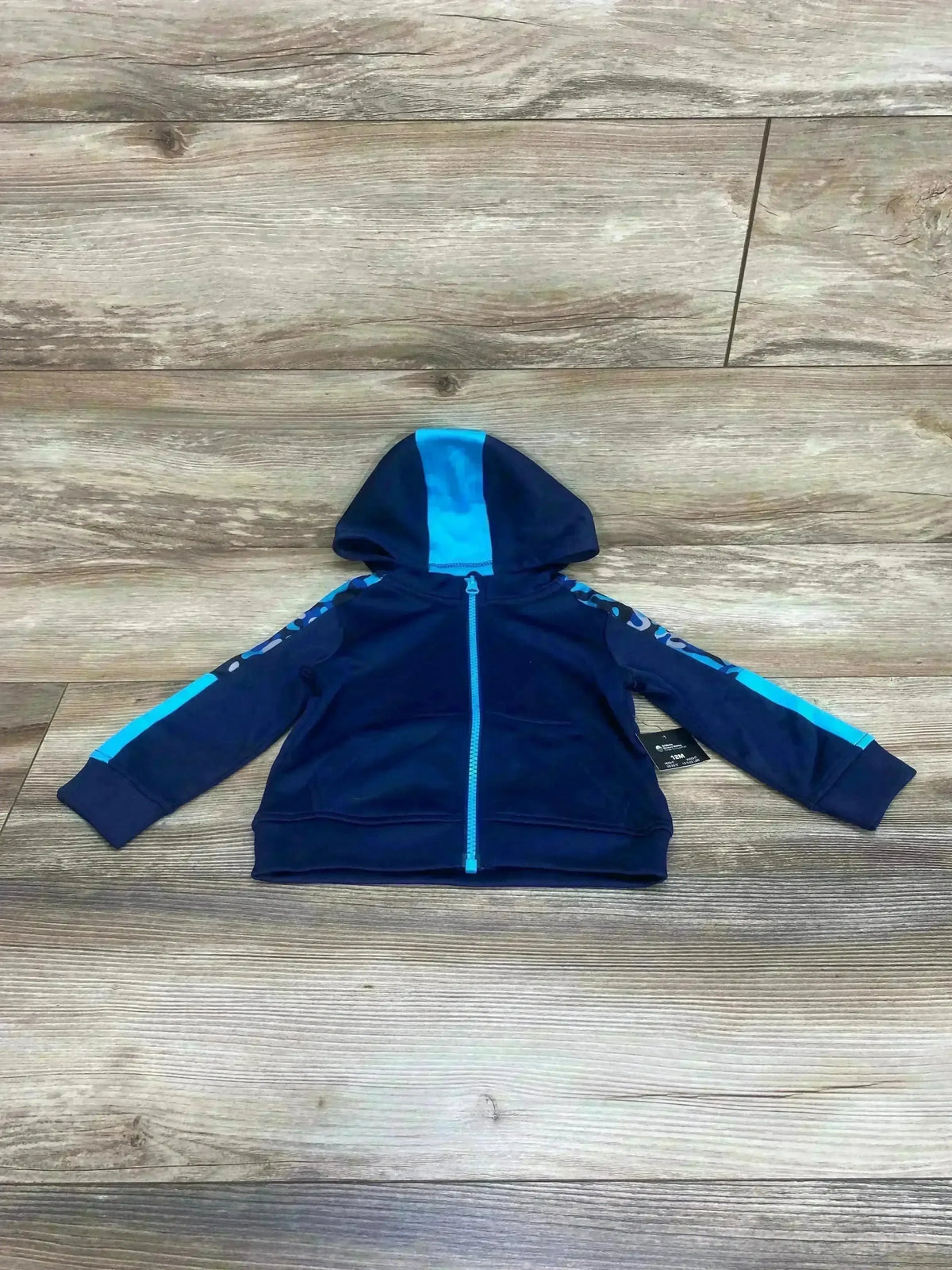 Top-down view of navy full-zip baby hoodie (12 mo) showing bright blue hood stripe, zipper, and sleeve stripes on wood floor