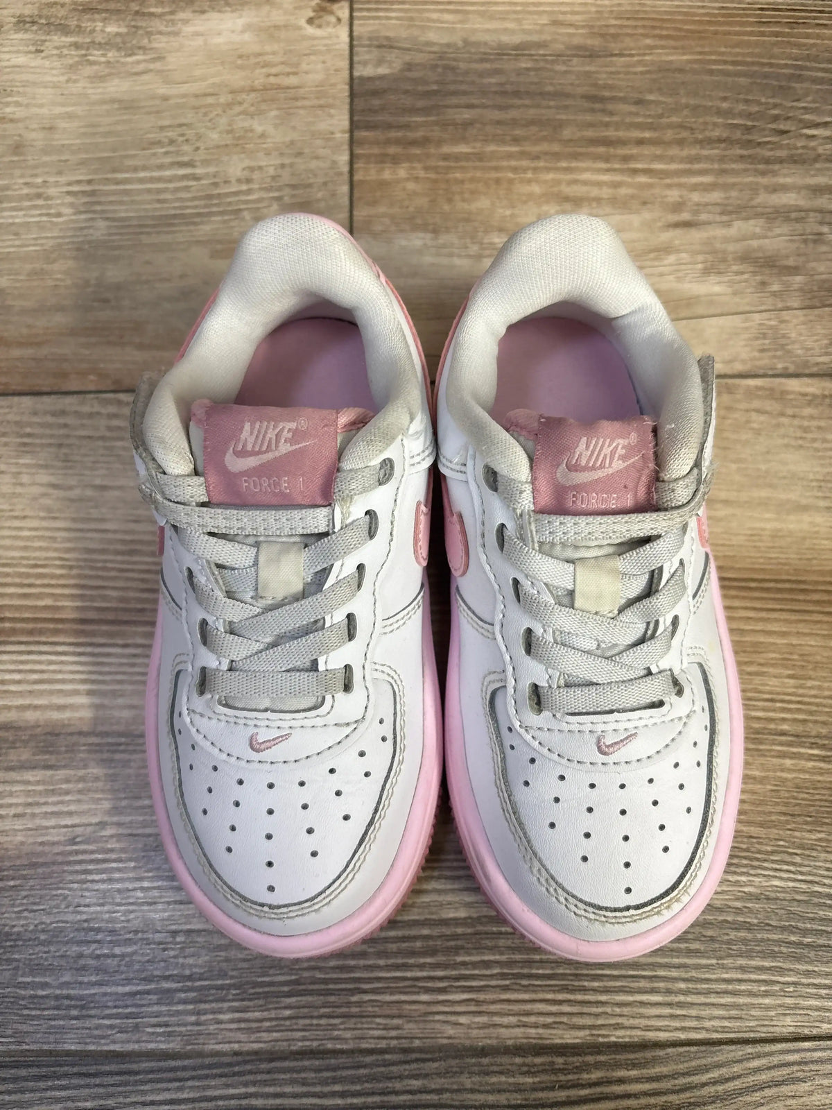 Top-down view of white toddler Nike Air Force 1 Low EasyOn sneakers showcasing pink midsoles, pink FORCE 1 tongue labels, perforated toes, and elastic laces on wood floor