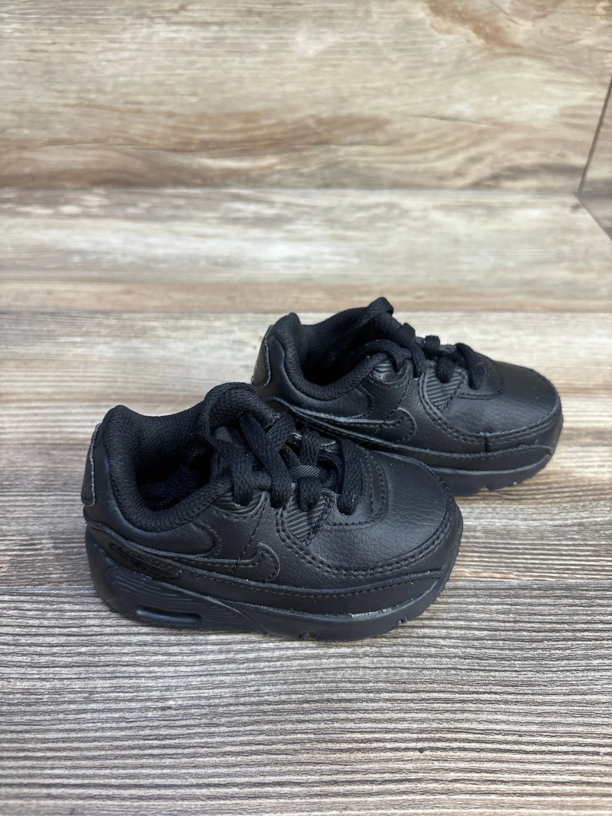 Three quarter view of Nike Air Max 90 TD sneakers showcasing the all black colorway on a wood grain surface
