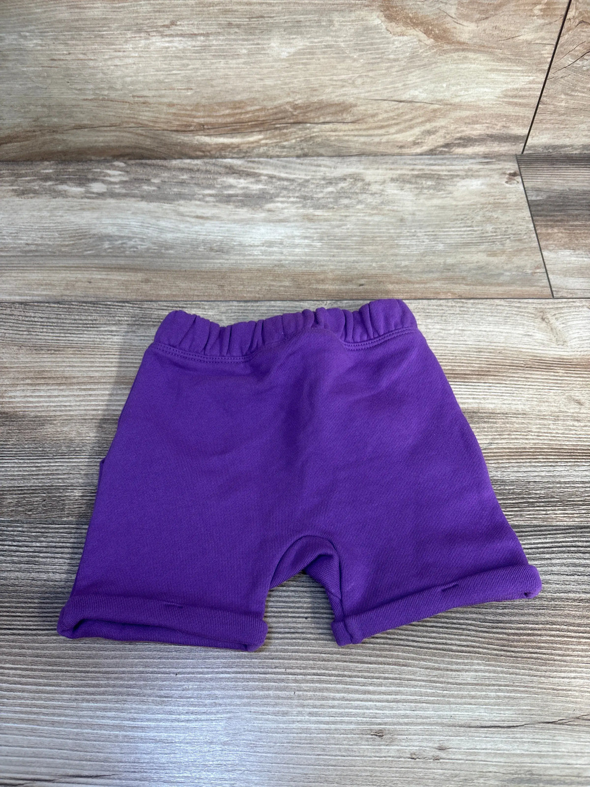 Top-down view of Disney pull-on purple newborn baby shorts showing elastic waistband and rolled cuffs on wood background