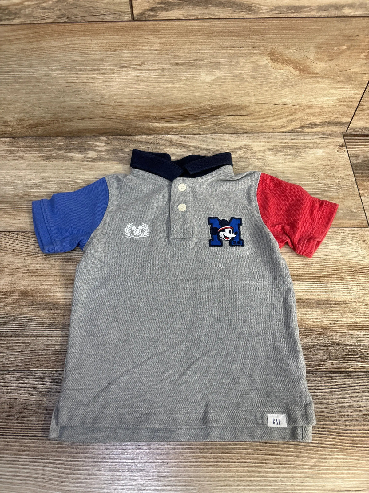 Front view of grey Baby Gap Mickey Mouse pique polo shirt with navy collar, colorful sleeves, and Mickey design on wood surface