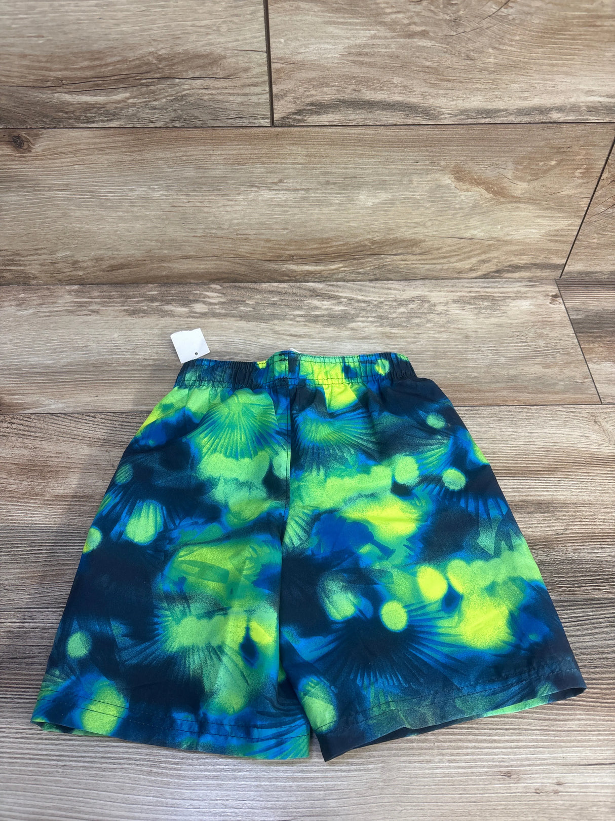 Front view of Under Armour swim trunks in size 4T showcasing a navy and bright green abstract pattern on a wood floor