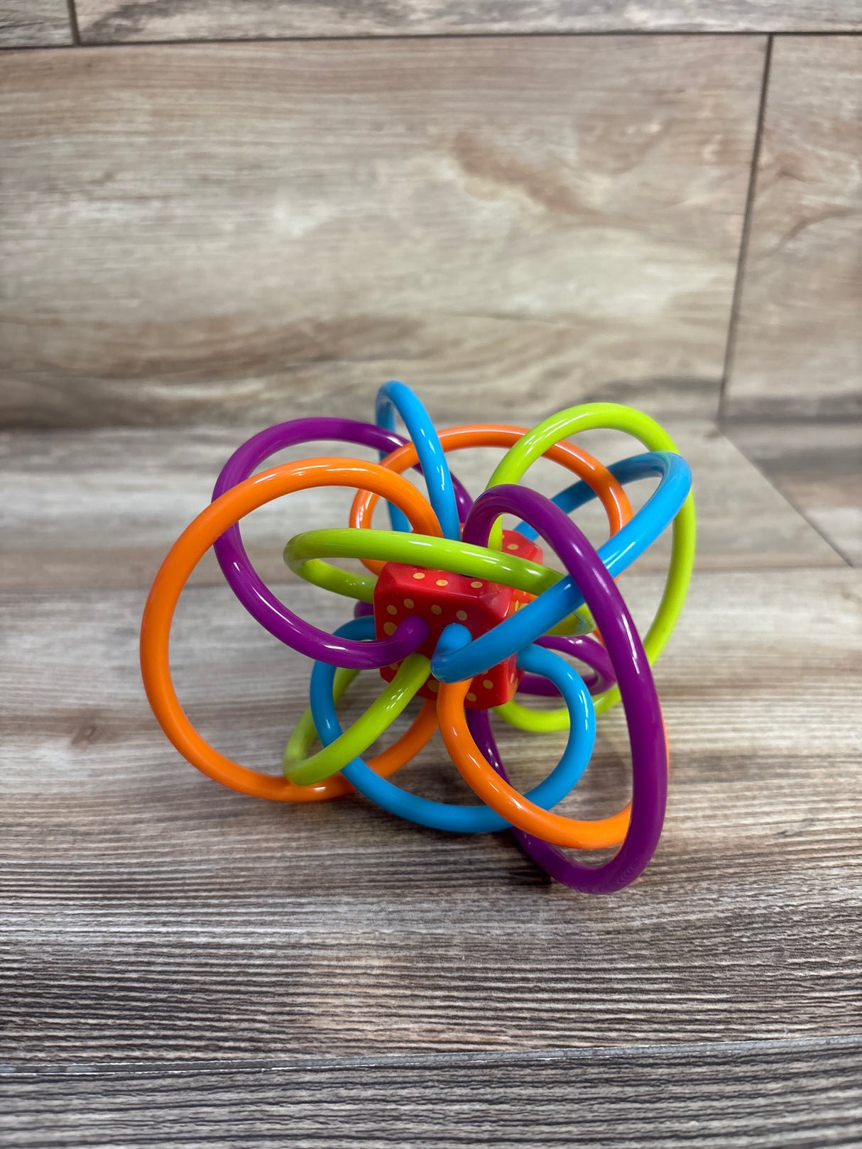 Angled top view of Winkel rattle and sensory teether showing multicolored interlocking rings and red dotted center on wood surface