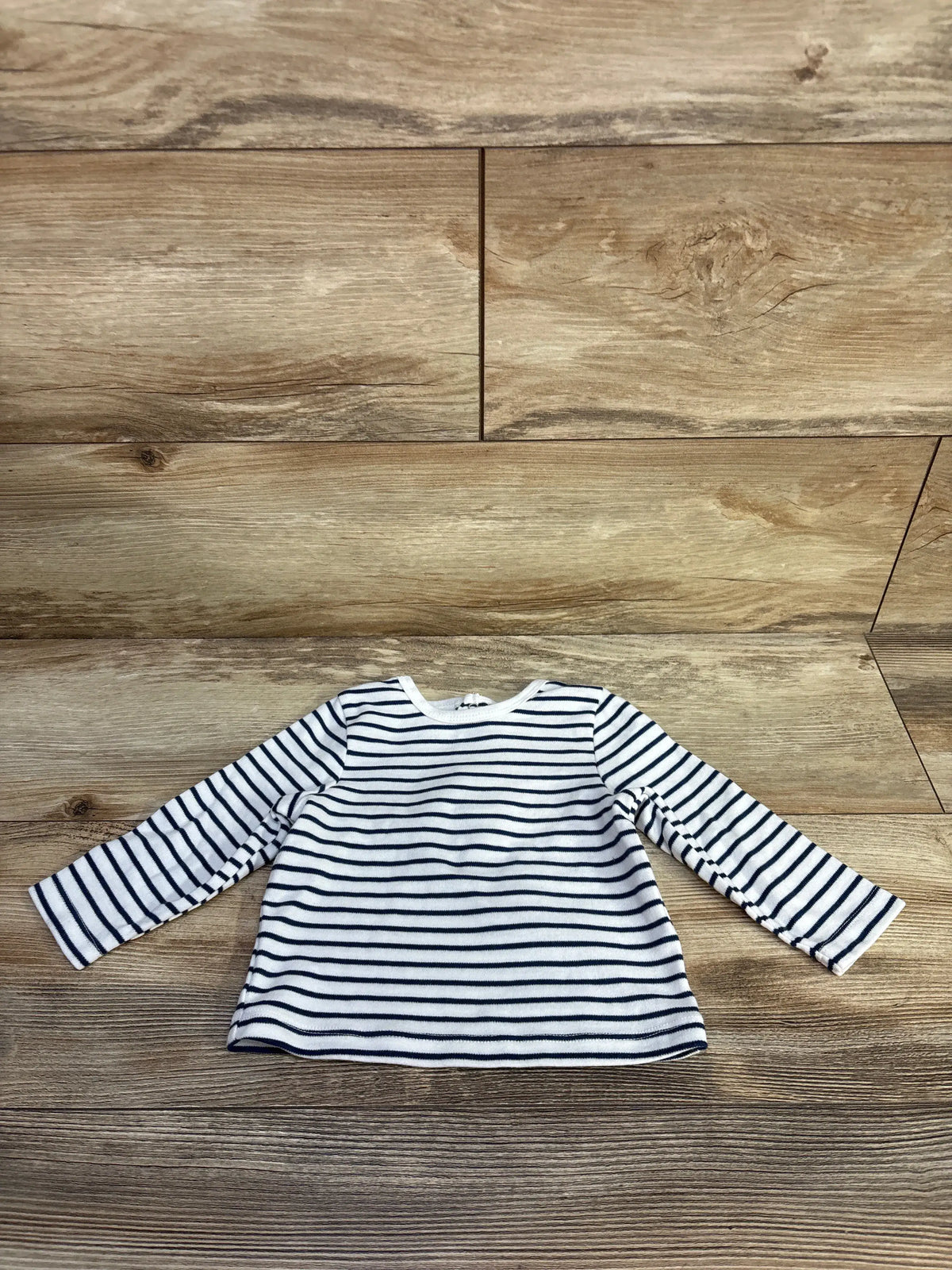 Front view of Little Me striped long sleeve shirt in white showcasing dark horizontal stripes with a wood floor background