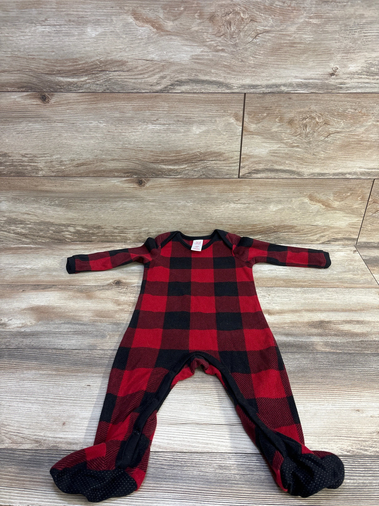 Top-down view of North Pole buffalo plaid blanket sleeper showcasing red and black check pattern, footed legs on light wood background