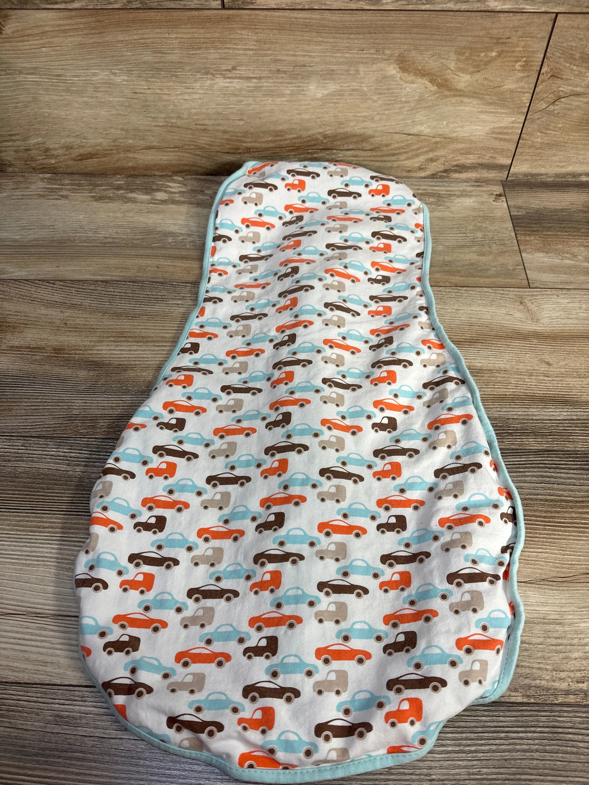 Top view of white Woombie Original Swaddle showcasing colorful car print and aqua edging on a wood floor
