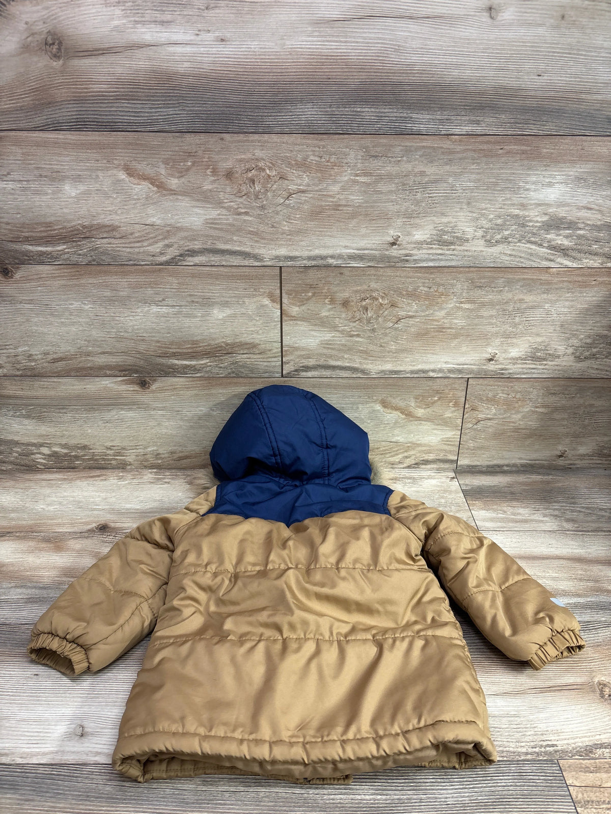 Back view of Oshkosh tan heavyweight ski jacket with navy hood and quilted sleeves on wood-grain flooring