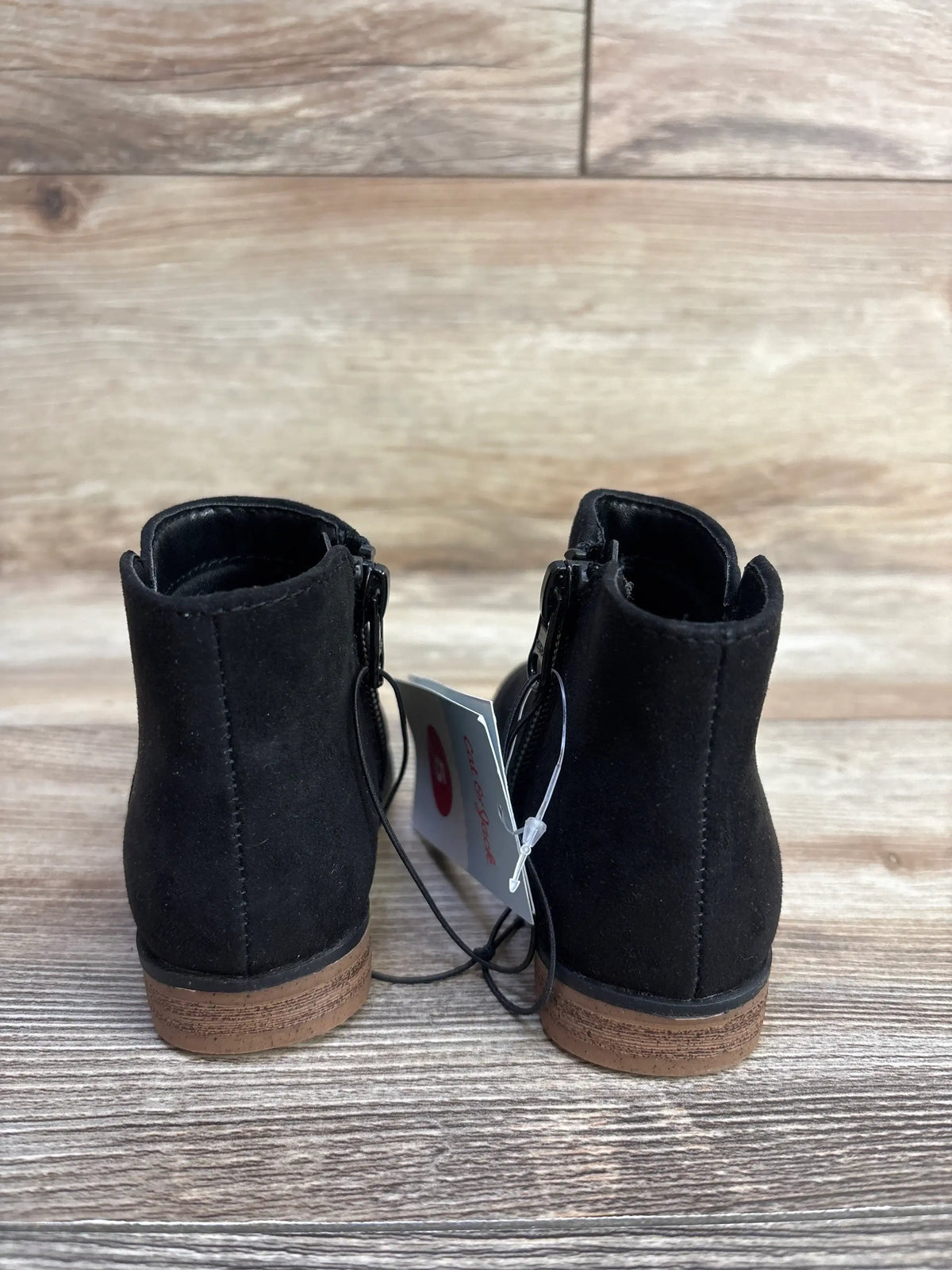Black suede ankle boots with brown soles on wood floor, rear view, product tag attached