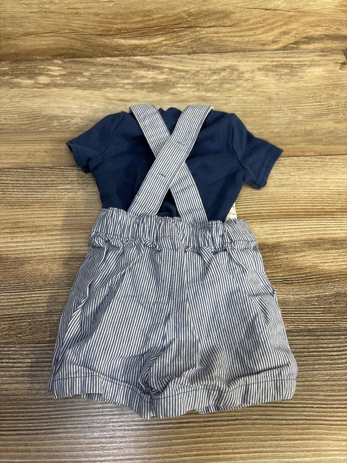 Front view of blue newborn 2-piece henley shirt and striped suspender shorts set with crossed suspenders on a wood surface