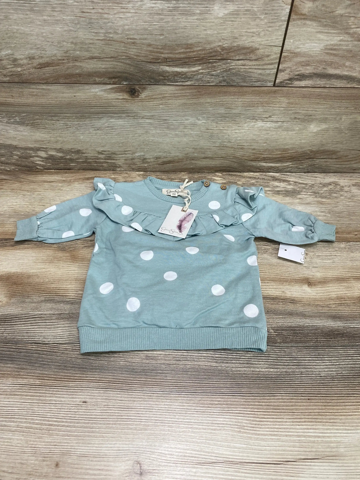 Flat lay of baby green polka dot sweatshirt dress showing ruffle yoke and shoulder buttons on light wood background