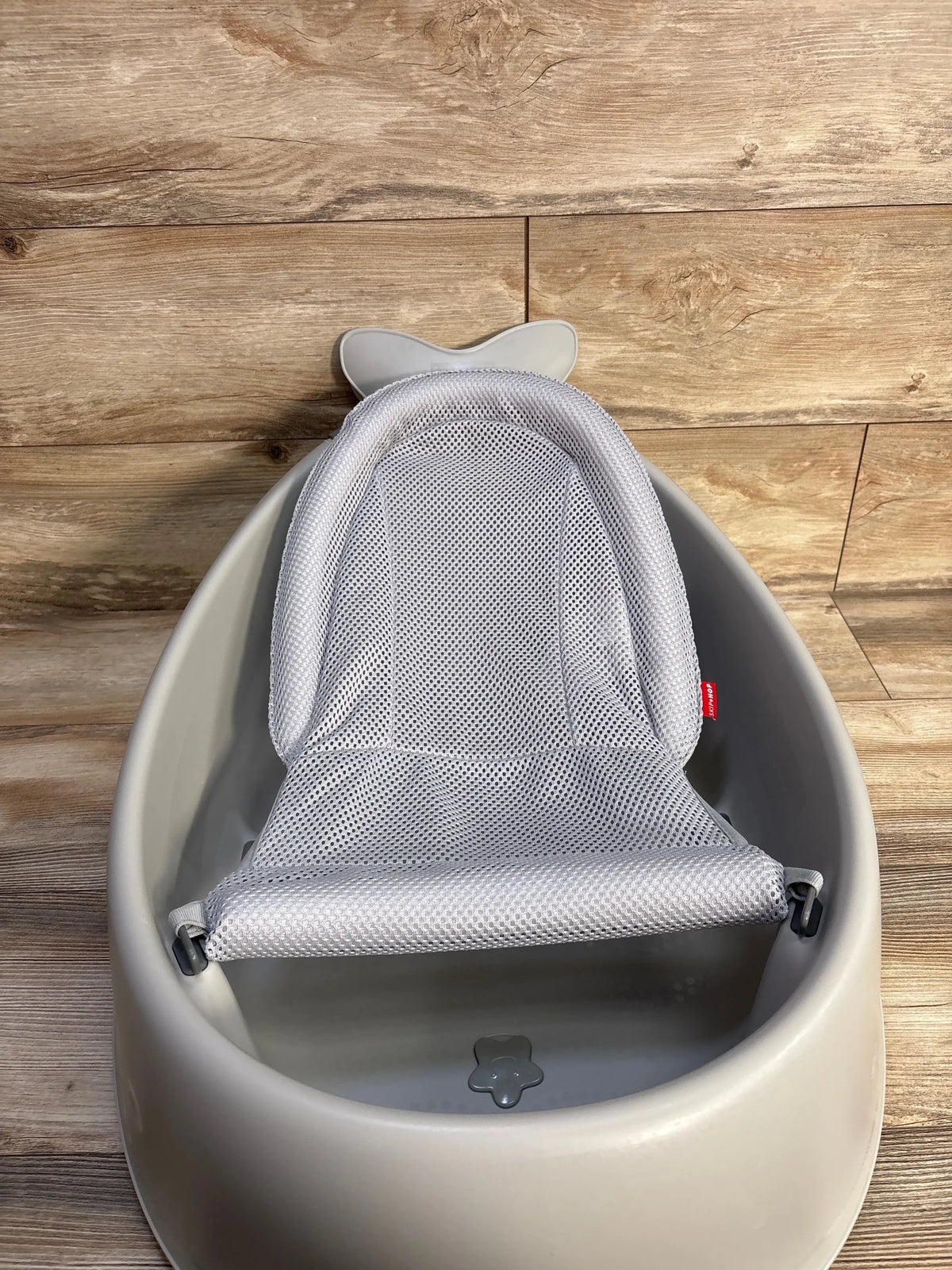 Top view of Skip Hop Moby Smart Sling 3 In 1 Baby Bathtub Grey showcasing mesh Smart Sling and drain plug on wood floor