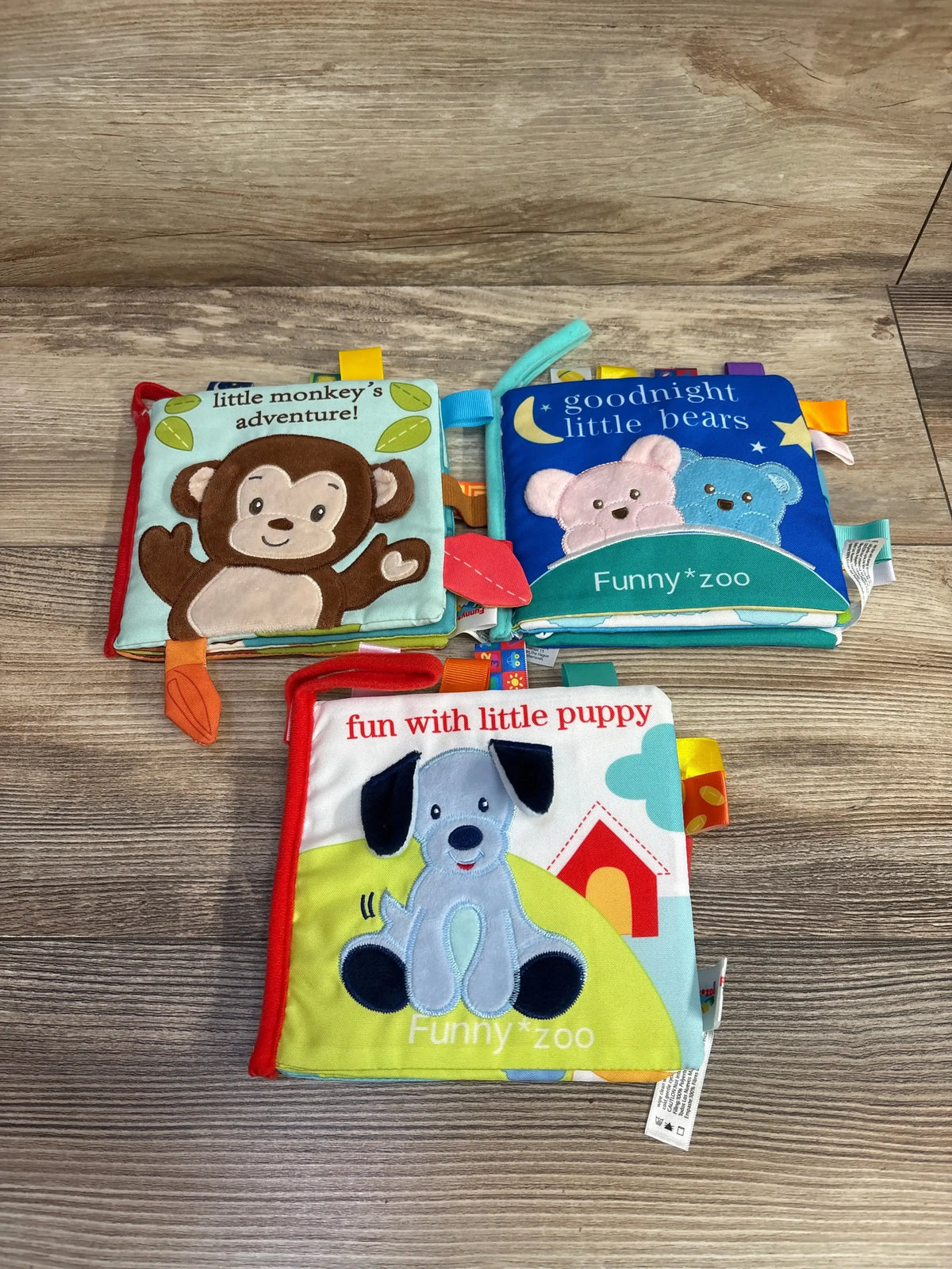 Three soft baby cloth books with animal covers on wood floor, featuring monkey, bears, and puppy.