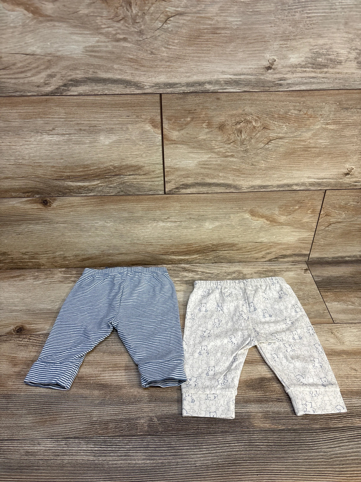 Flat lay of two Baby Gap pull-on newborn pants size 0-3M, blue striped pair and white pair with animal print on wood floor