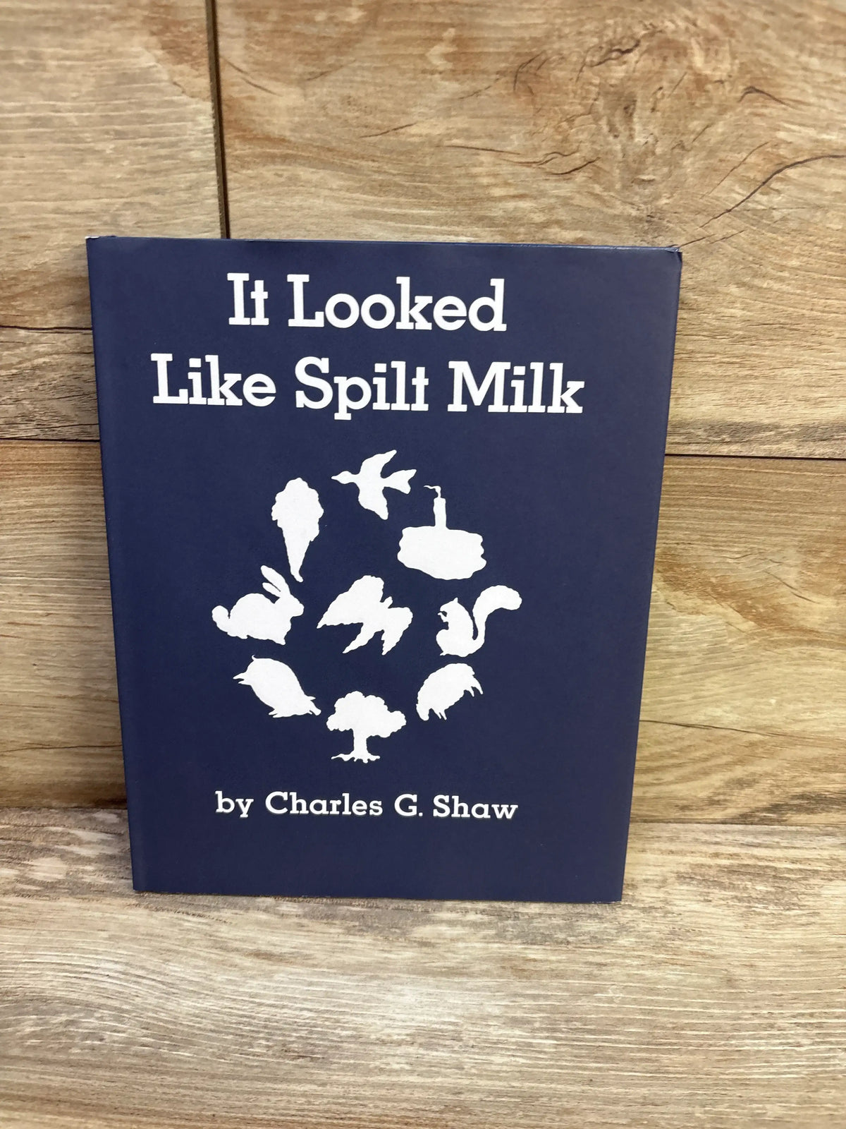 Front cover of It Looked Like Spilt Milk hardcover book showing white silhouette shapes and title on navy cover against wood background