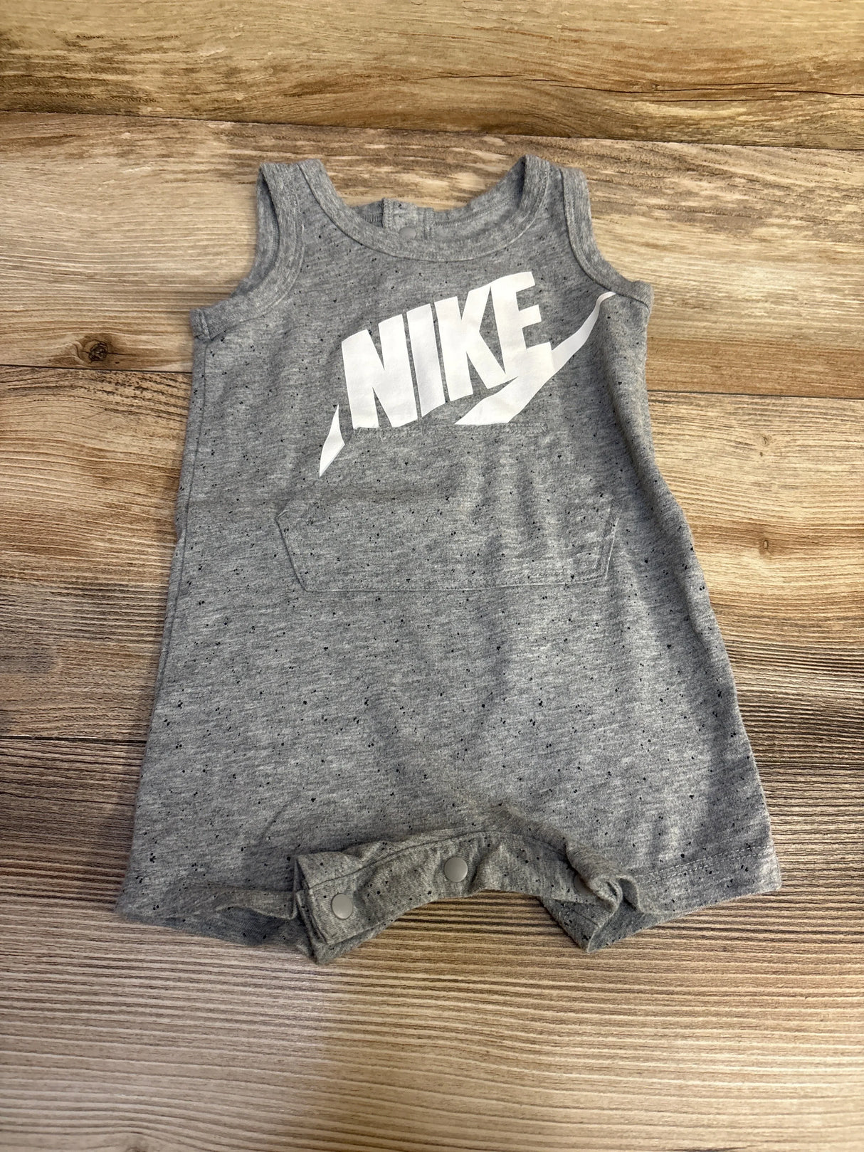 Flat lay of grey Nike sleeveless shortie romper with white NIKE logo, front pocket detail and crotch snap closures on wood
