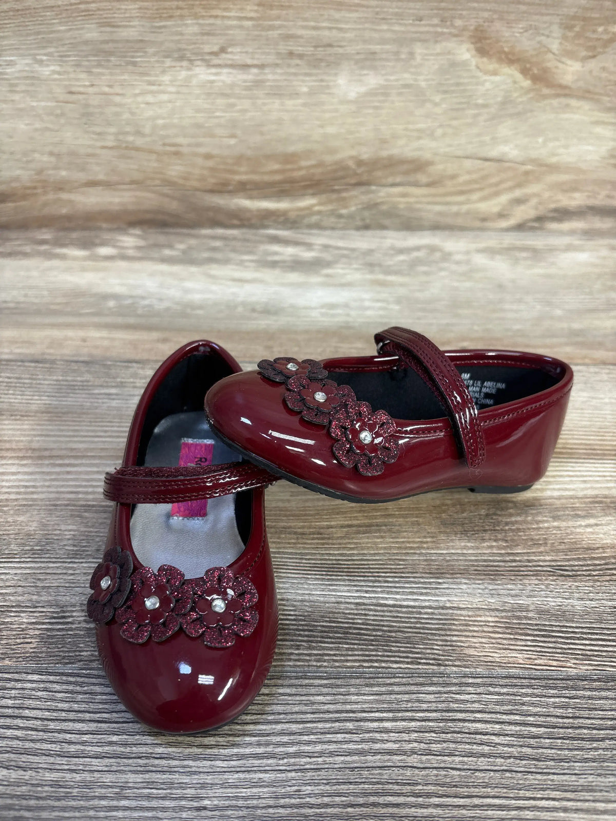 Top and side views of Rachel Shoes Lil Abelina flats with burgundy patent leather, floral accents, and halo straps