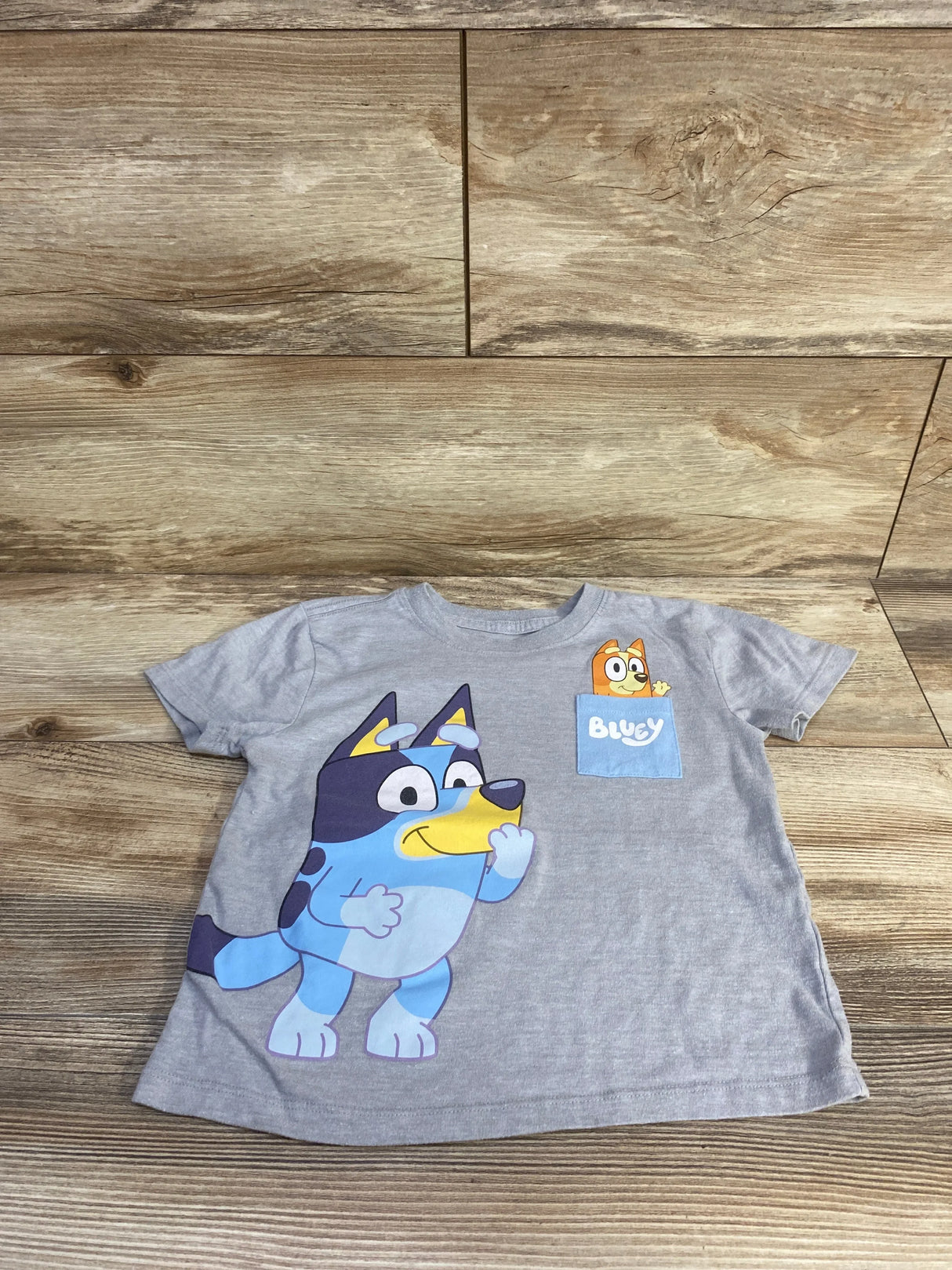 Flat lay of grey toddler T-shirt with large Bluey character print and a small pocket graphic labeled Bluey on wooden floor