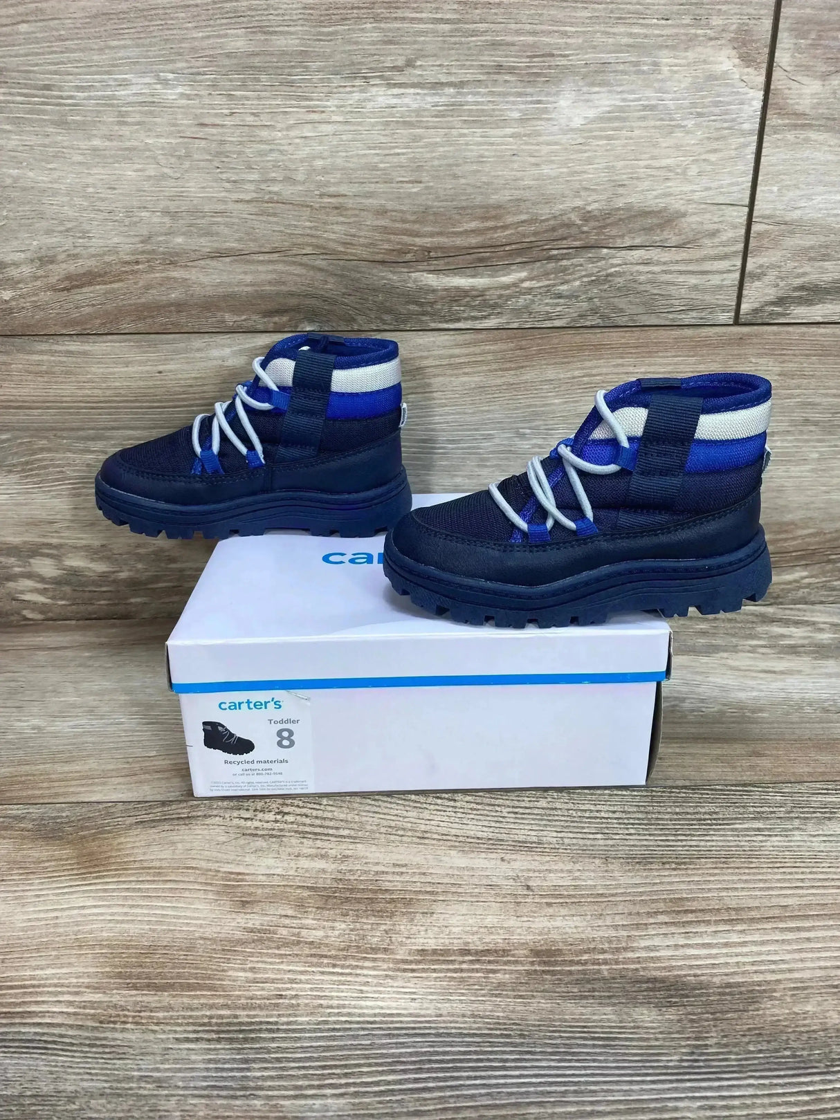 Side view of Carter’s toddler Fallon boots, navy blue with elastic laces and chunky tread on a shoebox with wood background