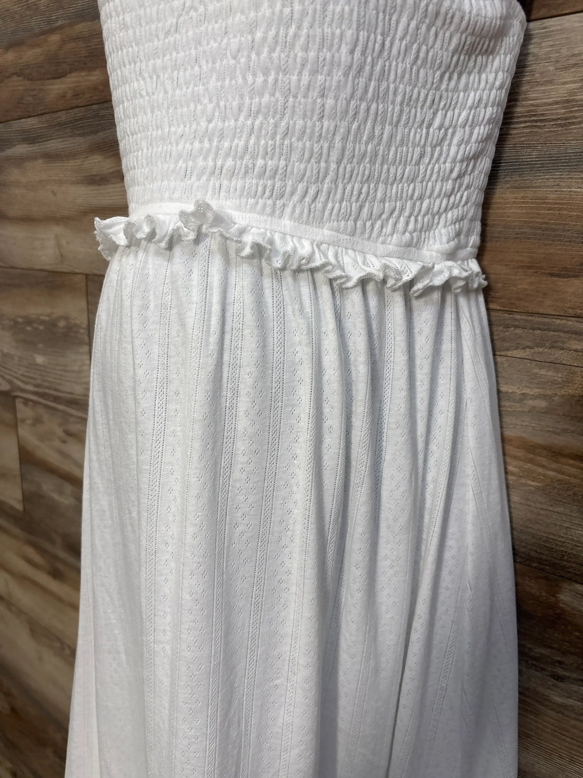 Close-up of white OUGES maternity boho maxi smocked dress showcasing textured smocked bodice and ruffled waist on a wood floor