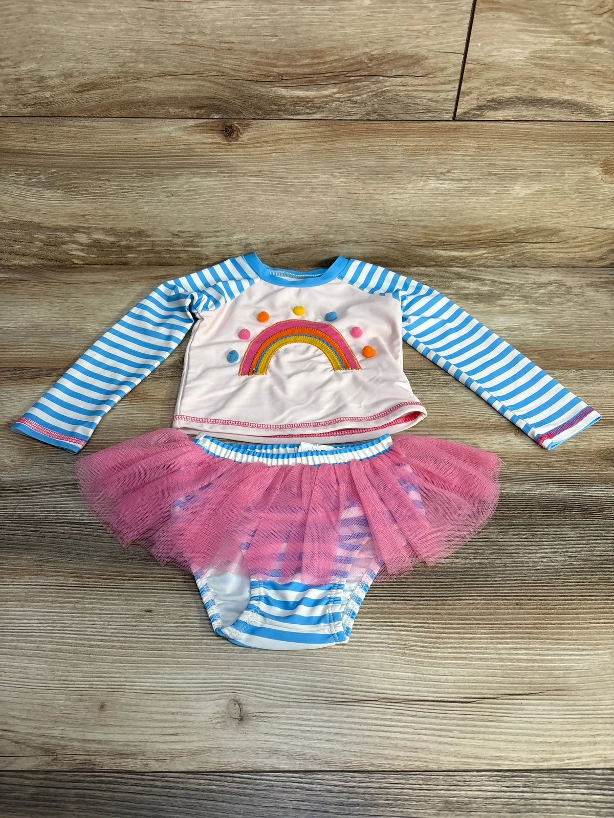 Flat lay of toddler rashguard swimsuit with rainbow embroidered top and striped bottoms with pink tulle on wood