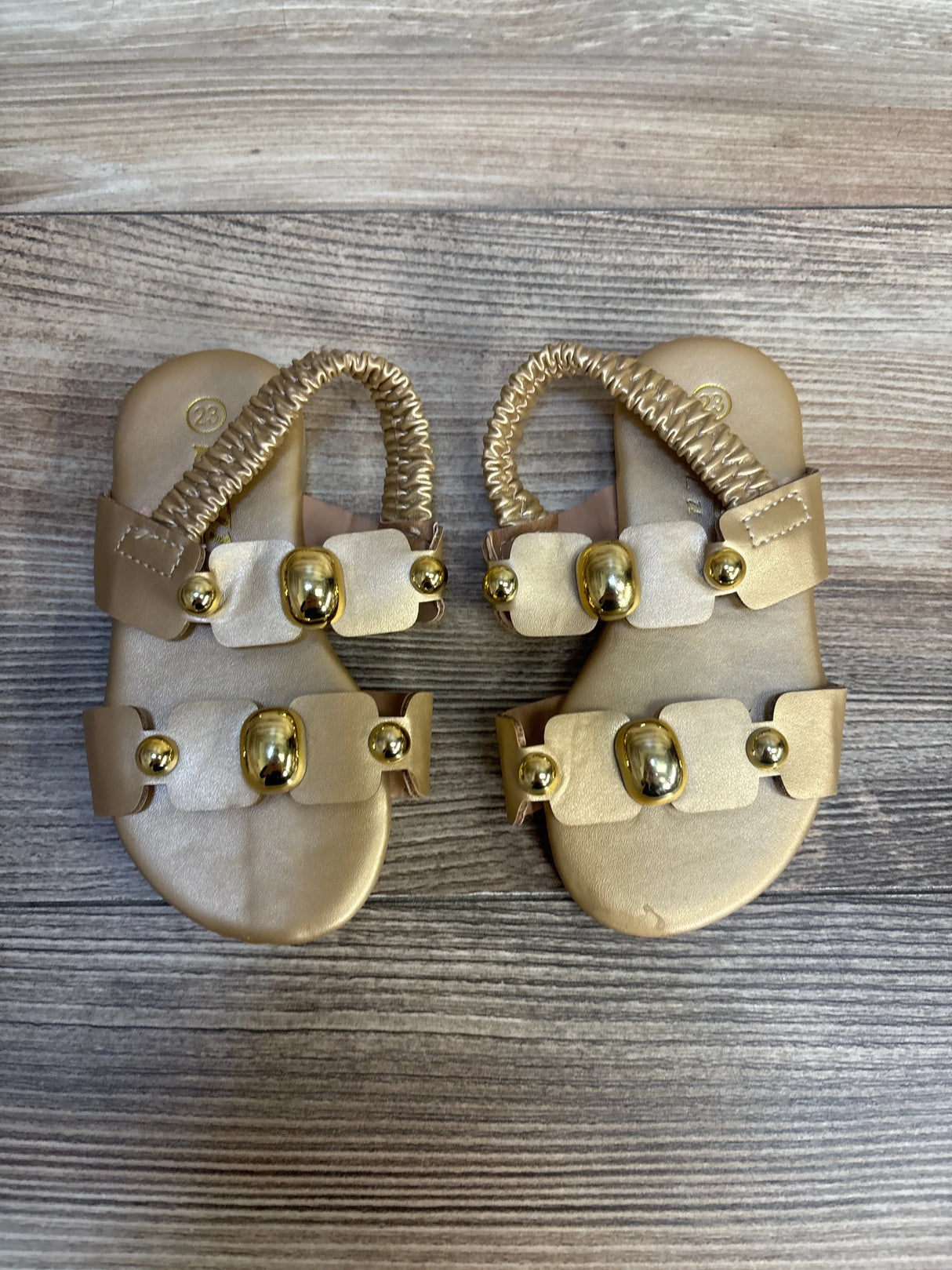 Gold toddler sandals with decorative beads and elastic ankle straps on wood floor