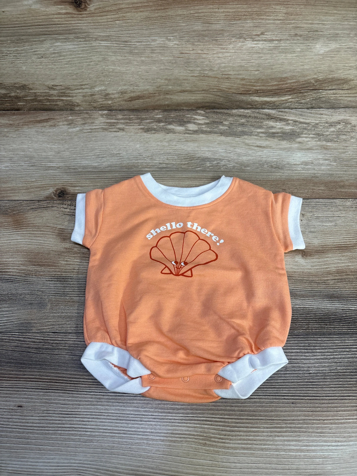 Flat lay of Cat and Jack terry romper in orange with Shello There seashell graphic and white trim on wood plank floor