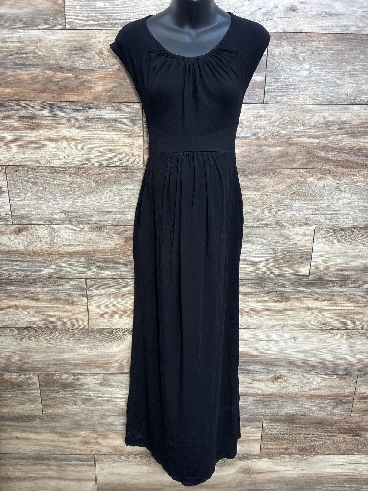 Front view of black maternity cap-sleeve maxi dress with empire waist and gathered skirt on mannequin against wood backdrop